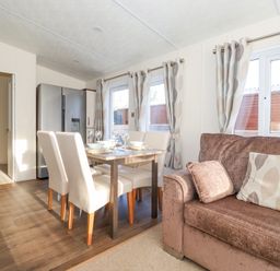 A dining area with a table set for four next to a brown sofa with cushions and windows with curtains at Draco in Felmoor Park
