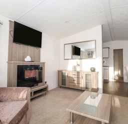 A living room with a sofa, coffee table, mounted TV, cabinet with mirror, and an open view into a dining area at Draco Felmoor Park