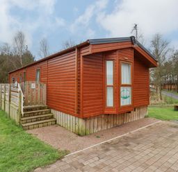 A wooden cabin with a small fenced porch and stairs surrounded by grass and trees at Ursa in Felmoor Park