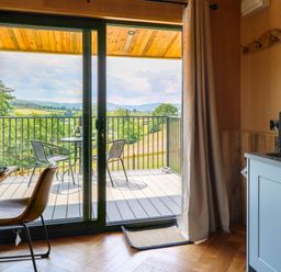 A kitchen area with blue cabinets and a sink next to a dining table with a chair by a sliding glass door opening to a balcony with two chairs and a table overlooking a landscape at Rooster's Rest in Rhaedr Gwy near Rhayader