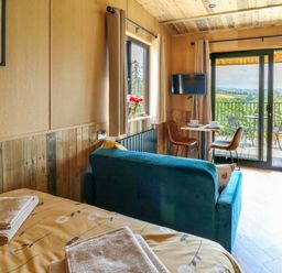 A room with a bed and towels a teal sofa a small dining table with two chairs a wall-mounted TV and a balcony overlooking a green landscape at Rooster's Rest in Rhaedr Gwy near Rhayader