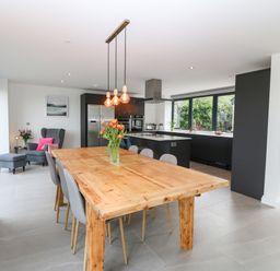 A dining area with a wooden table and grey chairs next to a kitchen with black cabinets and large windows at Pen-Y-Bryn in Llanfairpwllgwyngyll