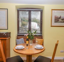 A dining area with a round wooden table set for two people near a window with blinds and framed artwork on walls at Bradley Stables in Withypool
