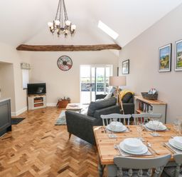 An open living and dining area with a wooden dining table set for four a gray sofa TV on a stand and a wooden beam on the ceiling at Pip's Barn in Brynteg near Benllech