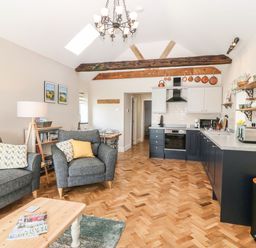 An open plan living area with grey armchairs and a sofa a wooden coffee table and a kitchen with black lower cabinets and cream upper cabinets at Pip's Barn in Brynteg near Benllech