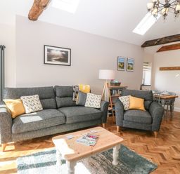 A living room with a gray sofa and armchair with yellow and patterned cushions a wooden coffee table a floor lamp and a dining table at Pips Barn in Brynteg near Benllech