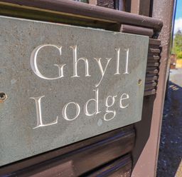 A close-up of a metal sign with the text Ghyll Lodge attached to a wooden post with a road and trees in the background at Ghyll Lodge Grasmere 25 White Cross Bay Windermere