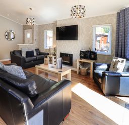 A living room with black leather sofas wooden tables a wall-mounted TV and windows at Ghyll Lodge Grasmere 25 White Cross Bay Windermere