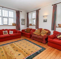 A seating area with four red sofas a patterned rug wooden floor and windows with curtains at Wick Court Farm in Arlingham near Frampton On Severn