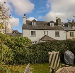 A garden with wooden chairs and a covered barbecue grill in front of a row of white and beige houses with slate roofs at Early Mist Cottage in Haytor Vale
