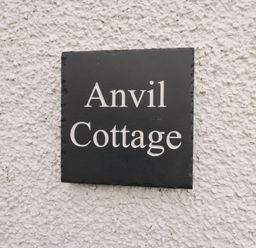 A black sign with white text reading Anvil Cottage on a rough white wall at The Anvil in Broughton-In-Furness