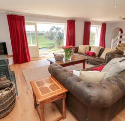 A living room with brown leather sofas a wooden coffee table flowers in a vase red curtains a wood stove and a basket of firewood at Whiteadder Bank in Berwick-Upon-Tweed
