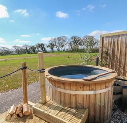 An outdoor hot tub made of wood with a metal stove and a wooden paddle on a deck with grass and trees in the background at Buzzards in Morchard Bishop