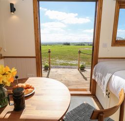 A small dining area with a wooden table and chair next to a bed and an open door leading to a green field at Buzzards in Morchard Bishop