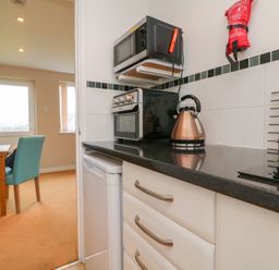 A kitchen counter with a kettle toaster microwave and fridge next to a dining area with blue chairs and wooden table at Sky Pocket in Bude