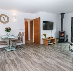 A dining area with a glass table and four chairs next to a living space with a TV mounted on the wall and a wood burning stove at Strawberry Bank - Nuthatch in Pyworthy near Bridgerule