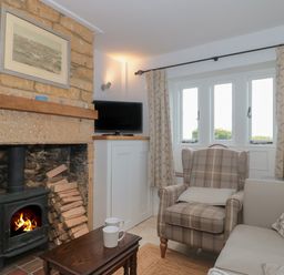 A sitting room with a wood burning stove and stacked firewood a plaid armchair a sofa windows with curtains and a small table with two mugs at Scrambles Cottage Stoke-Sub-Hamdon Somerset