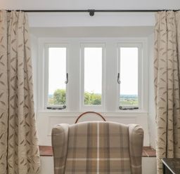 A window with three panes flanked by curtains and a plaid armchair facing the window at Scrambles Cottage in Stoke-Sub-Hamdon Somerset