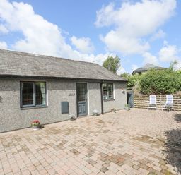 A courtyard with a paved ground and a gray building with two windows and a door with flower pots and two chairs at Cae Ffynnon Star near Gaerwen
