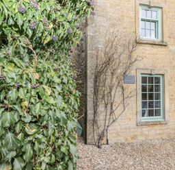 Side of a stone building with two windows and climbing plants next to a leafy shrub at Barton Cottage in Bourton-On-The-Water