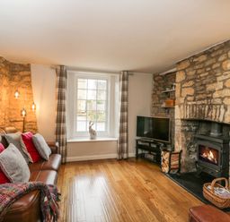 A living room with a brown leather sofa with cushions a window with plaid curtains a television and a stone fireplace with a wood burner at Barton Cottage in Bourton-On-The-Water