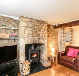 A living room with a stone fireplace wood logs a brown leather armchair with a pink cushion and a television on a stand at Barton Cottage in Bourton-On-The-Water