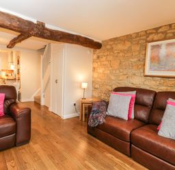 A living room with brown leather sofas with pink and gray cushions a stone wall with framed artwork a wooden floor and a lamp on a small table at Barton Cottage in Bourton-On-The-Water