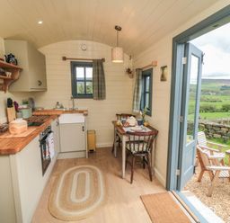 A small kitchen with wooden countertops a sink two chairs and a table next to an open door showing a countryside view at Sheep Cote Shepherds Hut in Chop Gate near Stokesley