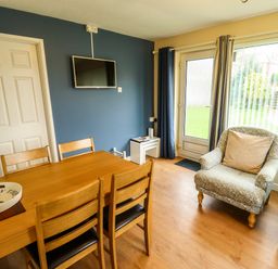 A dining area with wooden table and chairs a mounted TV an armchair near a window and door at 8 Royal Chalet Park in Mundesley