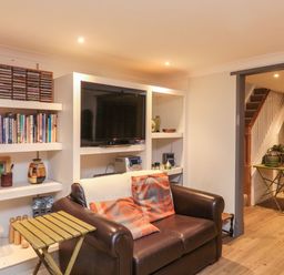 A living room with a brown leather sofa and orange pillows a TV in a white shelving unit books and CDs on shelves and a green side table with a potted plant at Lime Court Barn Sheepwash
