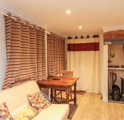 A room with a sofa and patterned cushions a wooden table with a chair curtains and a wood-burning stove at Lime Court Barn in Sheepwash