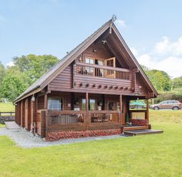 A wooden cabin with a balcony a porch and surrounding grass with trees at Robin Lodge in Llanbedr