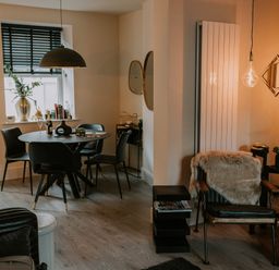 A dining area with a round table and four chairs next to a window with blinds and a living area with an armchair covered with a fur throw a small black side table and decorative lighting at 37 Jubilee Street in Llandudno