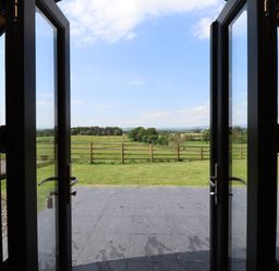 A view through open glass doors to a patio and green fields with a wooden fence at Cleugh Lodge in Lockerbie