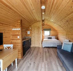 A wooden cabin interior with a bed at the far end a kitchen area a dining table with chairs a couch and a wall-mounted TV at Cleugh Lodge in Lockerbie