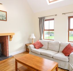 A living room with a beige sofa with red cushions a wooden coffee table a wood burning stove and two windows with curtains at Ballycahane in Castletownshend County Cork