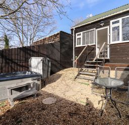 A small outdoor patio area with a hot tub a glass table with two chairs and stairs leading to a wooden house at Stillness in Blandford Forum