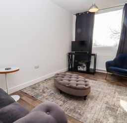 A living room with a gray sofa a blue armchair a gray ottoman a small round table a television on a black stand and gray curtains at Stillness in Blandford Forum