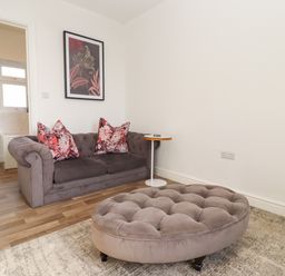 A living room with a gray sofa with floral pillows a round tufted ottoman and a side table next to an open door showing a bathroom at Stillness in Blandford Forum