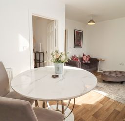 A room with a round white marble table and two chairs a grey sofa with cushions and a grey ottoman on a rug at Stillness in Blandford Forum