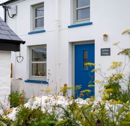 The exterior of a white cottage with a blue door and two windows with blue trim and a garden in the foreground at Dyffryn Cottage in Aberporth