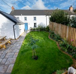 A backyard with a green lawn two small trees a wooden picnic table and a paved walkway next to a white house with a blue door at Dyffryn Cottage in Aberporth