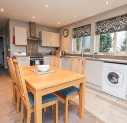 A kitchen with wooden dining table and chairs washing machine oven microwave and windows with patterned blinds at Beech Lodge in Goosnargh