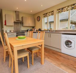 A kitchen with a wooden dining table and four chairs a washing machine under the counter and three windows with patterned blinds at Oak Lodge in Goosnargh