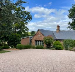 A stone house with a slate roof surrounded by trees and bushes with a gravel driveway at Court Lodge in Bathealton near Wiveliscombe