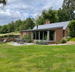 A house with outdoor dining furniture and a lounge chair on a grass lawn with trees in the background at Court Lodge in Bathealton near Wiveliscombe