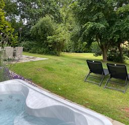An outdoor garden area with a hot tub in the foreground two black lounge chairs on grass trees and a stone patio with a dining table and chairs at Court Lodge in Bathealton near Wiveliscombe