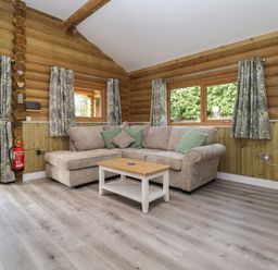 A living room with a beige sectional sofa green patterned cushions a wooden coffee table wood-paneled walls and windows with leafy curtains at Redwood Lodge in Ellesmere