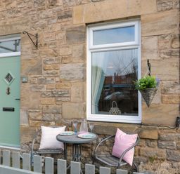 A small outdoor seating area with two chairs and a round table set against a stone wall with a green door and a window with a hanging plant at Cosy Cottage in Amble