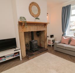 A living room with a wood stove fireplace a gray sofa with pink cushions a white TV stand with a television and board games at Cosy Cottage in Amble
